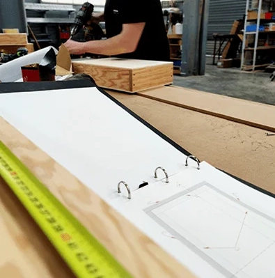 Bouw van een privébioscoopzaal waar een ambachtsman werkt aan de constructie van een houten element. Op de voorgrond ziet men een geopende map met technische plannen en een rolmaat op de werkbank